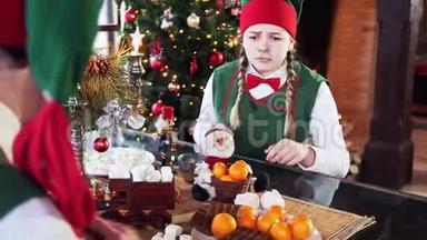 两个有趣的精灵坐在圣诞背景的桌子上。 女孩精灵试图带走糖果，另一个精灵干扰了她。